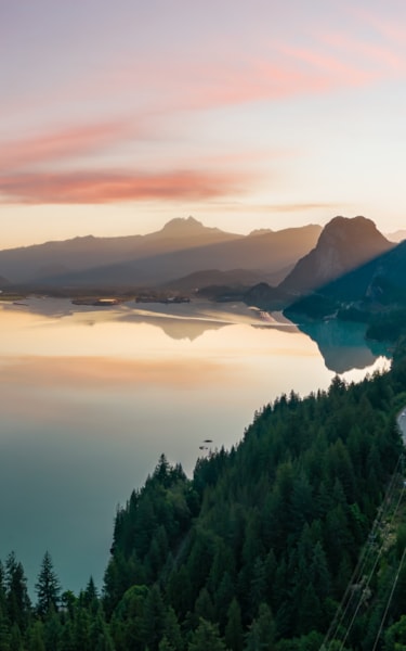 Stille innsjø som speiler fjell og himmel ved soloppgang eller solnedgang, med vei langs en skogkledd skråning.